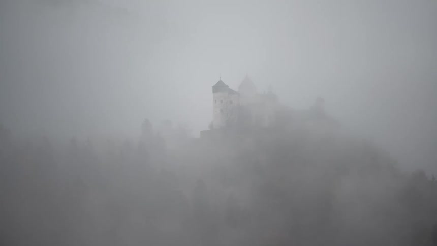 A mysterious medieval castle slowly emerges from the thick, swirling morning fog on a forested hillside