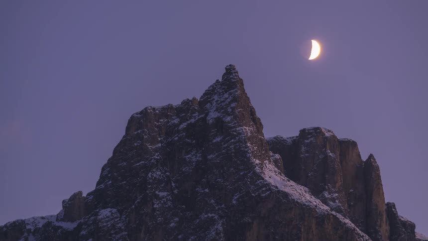 Timelapse of a crescent moon setting behind a majestic snow-covered mountain peak as the sky transitions from purple dusk to starry night
