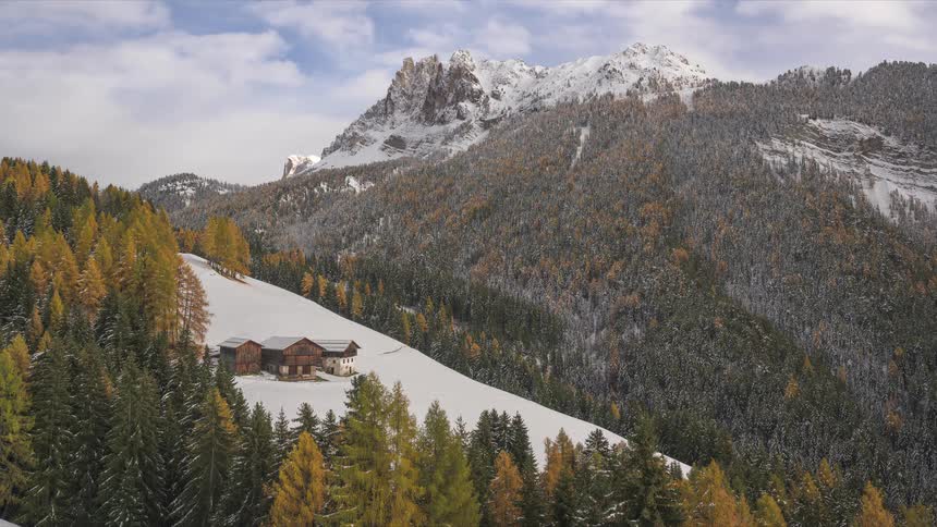 A timelapse captures moving clouds and sunlight over a snowy mountain landscape with autumn forests and a secluded farm in the Italian Dolomites