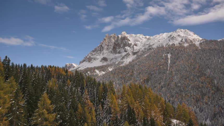 A timelapse captures clouds moving over a snow-capped mountain peak rising above a colorful forest of pine and golden larch trees