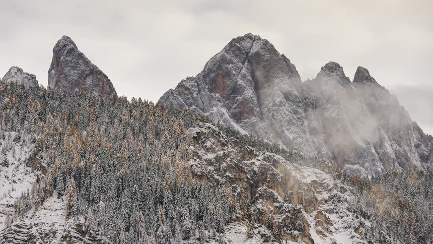 A stunning timelapse of clouds and fog moving over the majestic, snow-covered Dolomite mountains and a vast pine forest