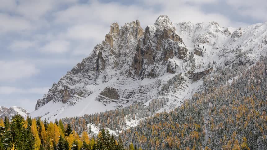 Clouds drift across a rugged, snow-covered mountain with a vibrant autumn forest at its base in a beautiful time-lapse