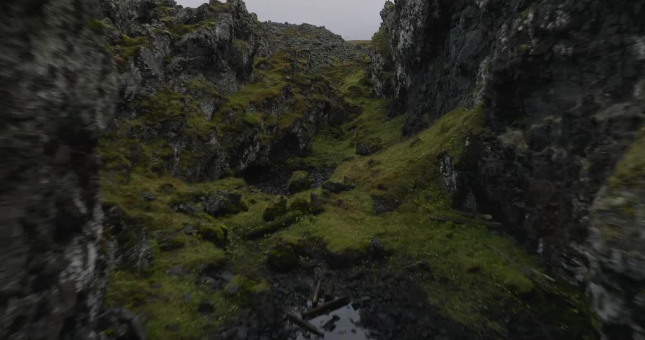 A dramatic aerial descent into a rugged, moss-covered volcanic rock canyon under an overcast sky