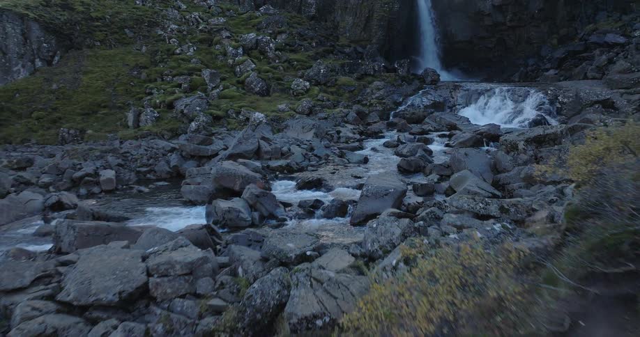 Following a person in a colorful jacket as they hike along a rocky trail towards a cascading waterfall
