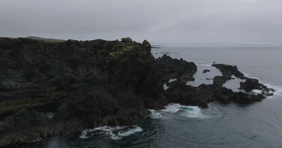 An aerial view pulls back from a mossy sea stack to reveal the dramatic and rugged volcanic coastline of Iceland under a cloudy sky