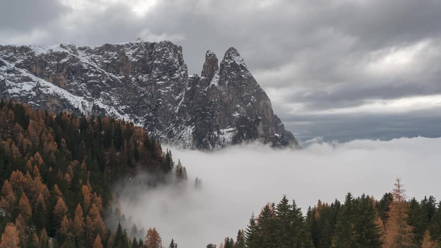A moody timelapse captures rolling fog and clouds engulfing a colorful alpine forest below towering, snow-covered mountain peaks