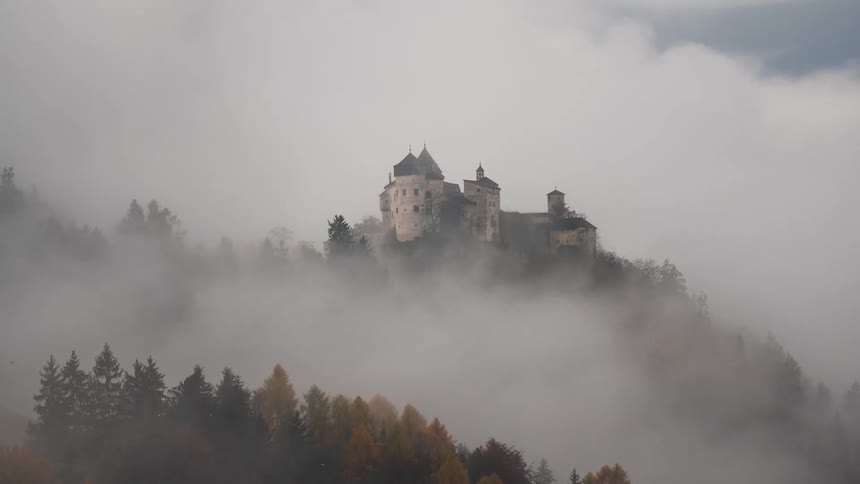 A mysterious medieval castle on a forested mountain peak is revealed and then concealed by thick, rolling fog in a dramatic time-lapse