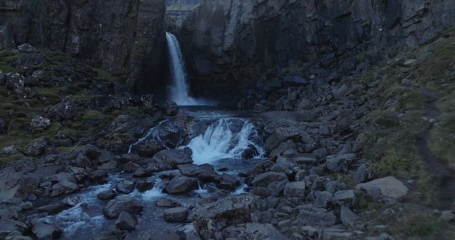 A dramatic aerial shot pushes slowly toward a powerful waterfall cascading into a rocky river inside a dark, secluded canyon