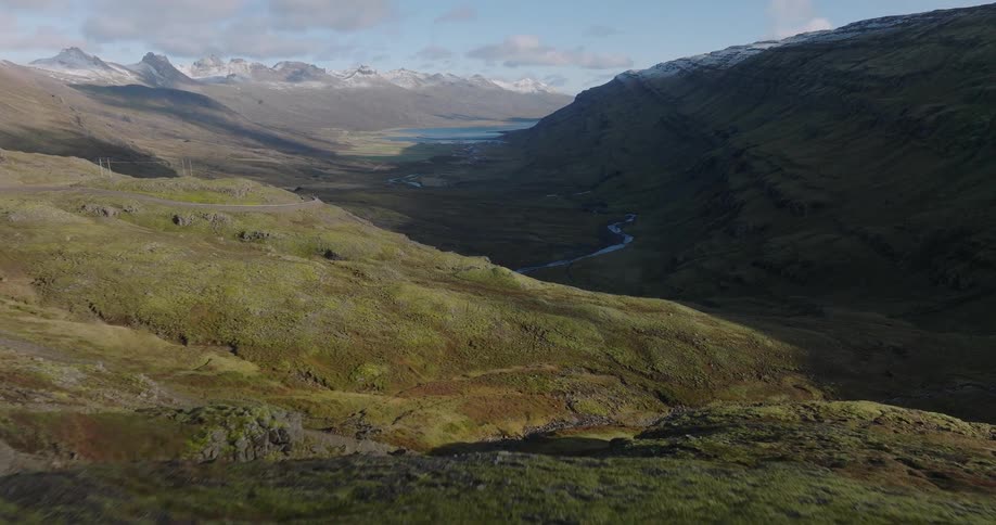 An expansive aerial shot flies over a green, mossy valley towards a winding river and distant snow-capped mountains
