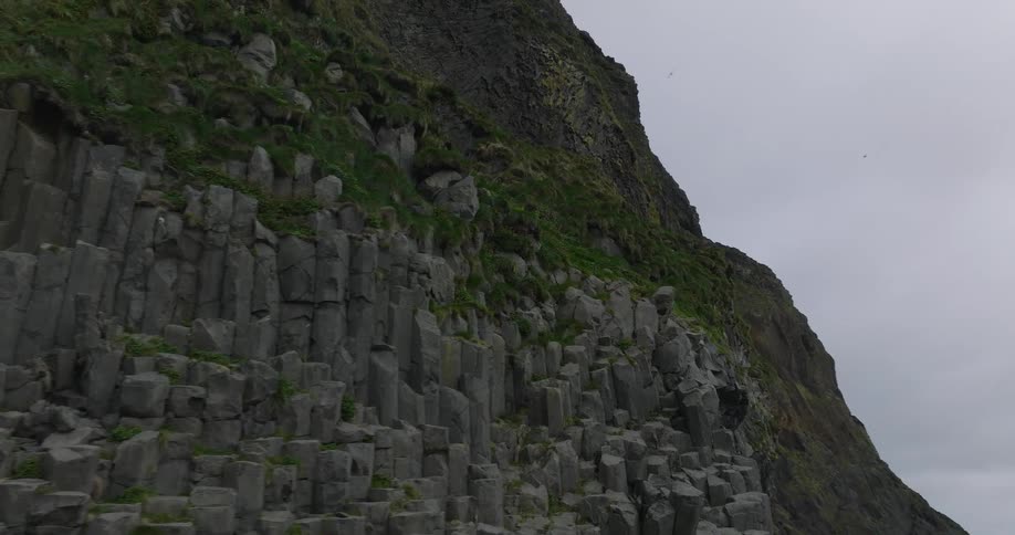 A slow aerial push in towards the majestic green-topped basalt columns of a coastal cliff in Iceland