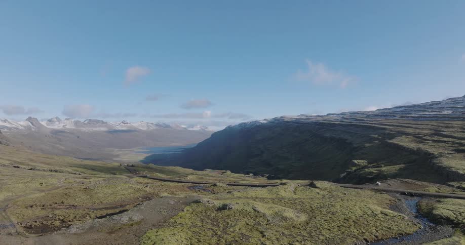 Expansive aerial pullback shot reveals a mossy green valley, a winding road, and snow-dusted mountains in Iceland