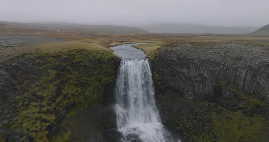 A dramatic aerial shot flies towards and over a majestic waterfall in the foggy, rugged highlands of Iceland