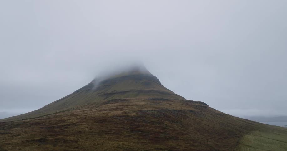 An aerial view pushes in on Iceland's iconic Kirkjufell mountain as its peak is enveloped by thick, moody clouds