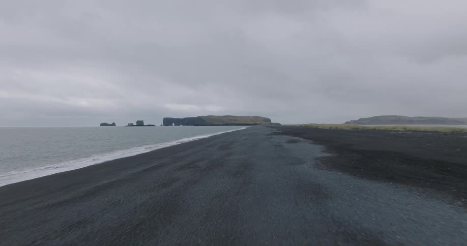 An immersive, low-level drone shot glides quickly over the volcanic black sand beach of Reynisfjara, Iceland on a gray, overcast day