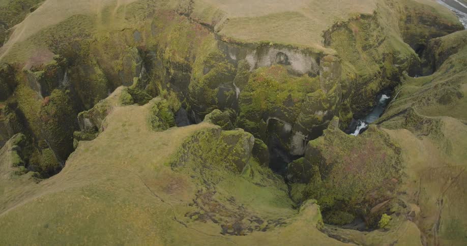 An epic aerial shot rotates over a vast, mossy green canyon carved by a winding river in Iceland