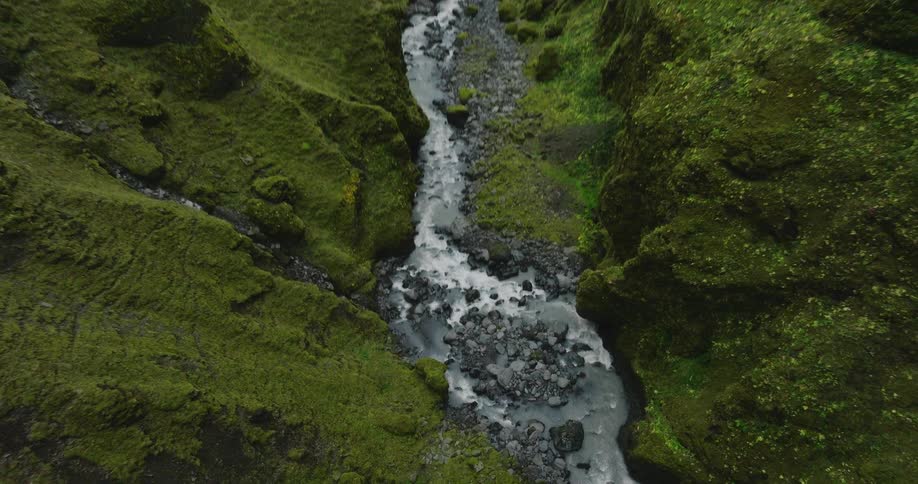 A stunning aerial shot tilting down on a river flowing through a deep, dramatic canyon covered in vibrant green moss in Iceland