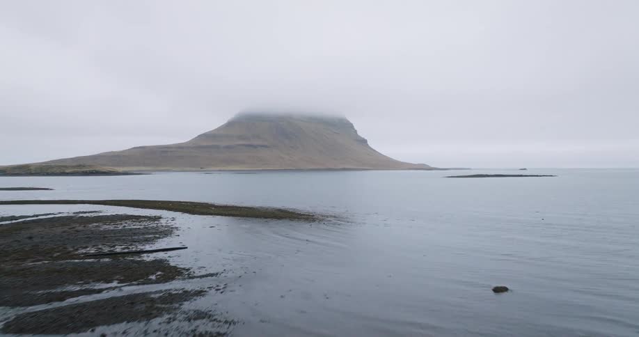 Aerial flight from the grassy shoreline over the calm water towards the famous Kirkjufell mountain in Iceland on a foggy day