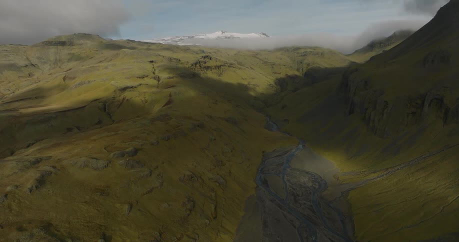 Sunlight and shadows from clouds move across a vast, green mountain valley where a river flows towards a distant snow-capped peak