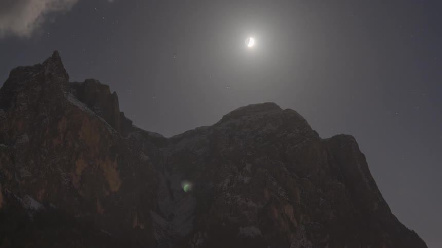 Time-lapse of a crescent moon and stars traveling across the night sky above a snowy mountain peak