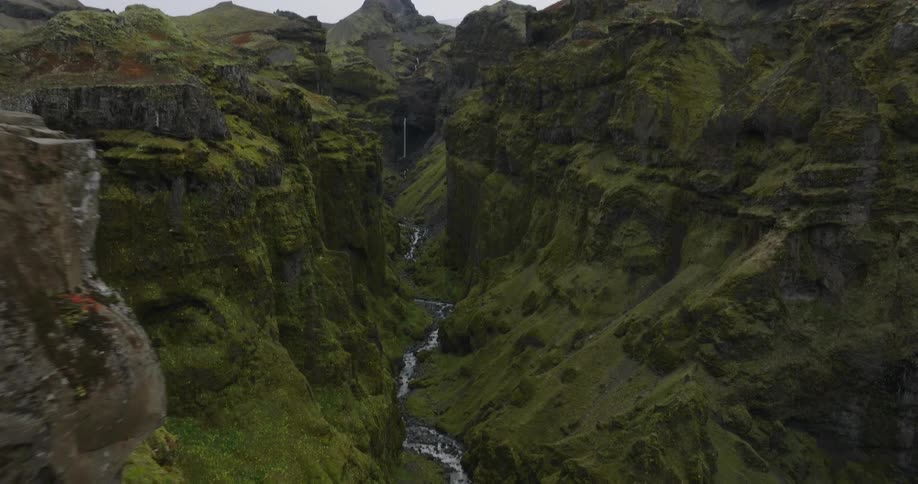 An aerial shot pushes past a woman in a flowing gown sitting on a dramatic cliff edge, revealing a majestic green moss-covered canyon with a winding river
