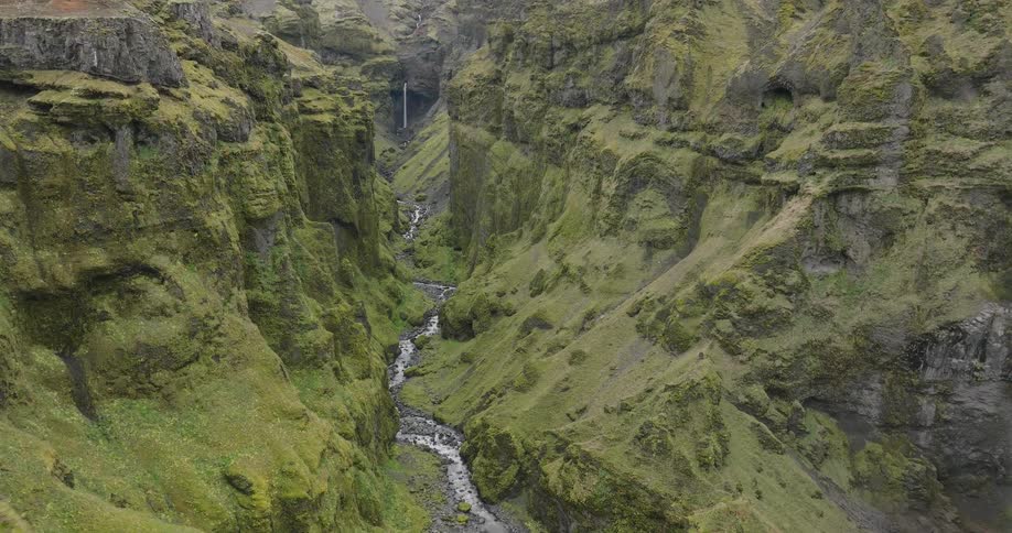 An aerial view pushes forward over the edge of a vast, moss-covered green canyon with a river and a distant waterfall