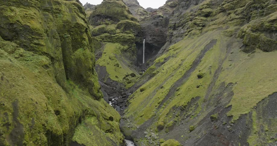 An aerial view reveals a stunning waterfall nestled deep within a lush, moss-covered canyon in the rugged landscapes of Iceland