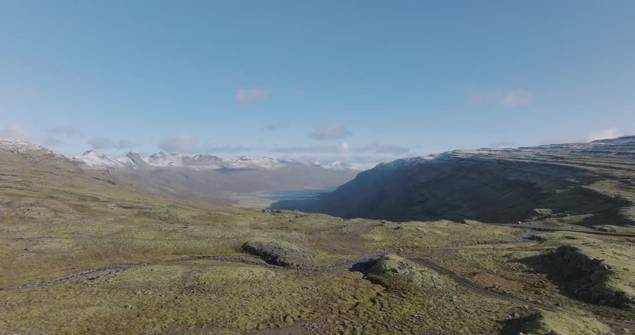 A slow aerial push-in shot over a vast, green moss-covered valley in Iceland, revealing a river and distant snow-capped mountains under a clear blue sky