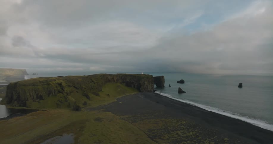 An epic aerial view reveals the stunning Dyrhólaey peninsula, with its famous rock arch and lighthouse, overlooking the black sand beaches of southern Iceland