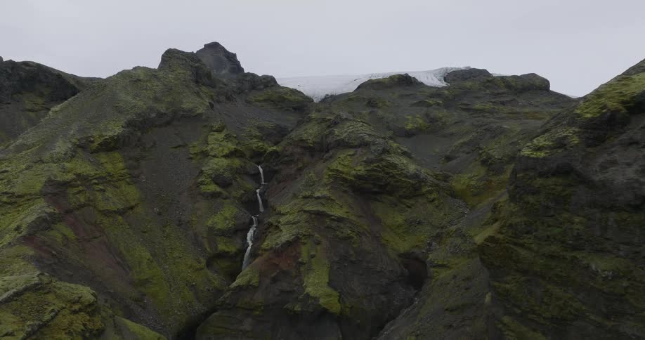 A dramatic aerial shot reveals a stunning waterfall cascading down a mosscovered volcanic mountain with a glacier on top in Iceland