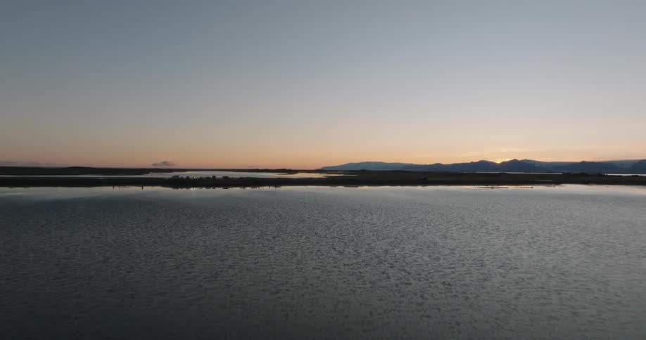 A tranquil aerial shot flies low over calm, reflective waters towards a distant spit of land at sunset in Iceland