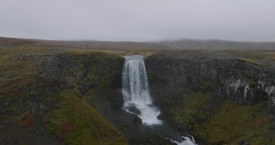 An aerial shot flies backward, revealing a powerful waterfall cascading over a cliff in a vast, desolate landscape on an overcast day