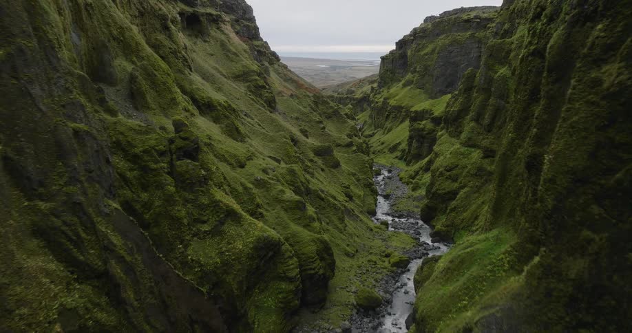 A majestic aerial dolly shot slowly reveals a stunning, lush green mossy canyon with a river winding through its rocky base
