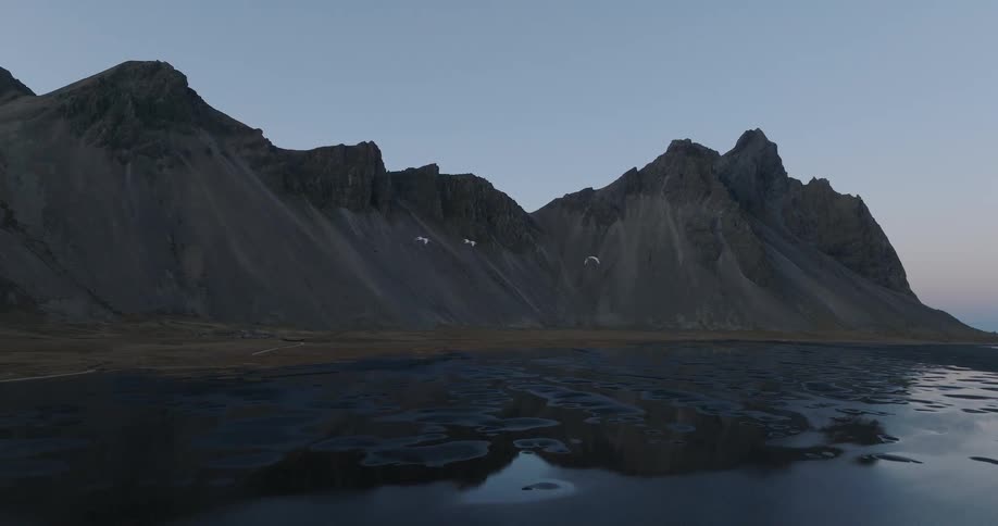 Slow motion aerial shot of white swans flying over a black sand beach with dramatic Icelandic mountains reflecting in the still water at dusk