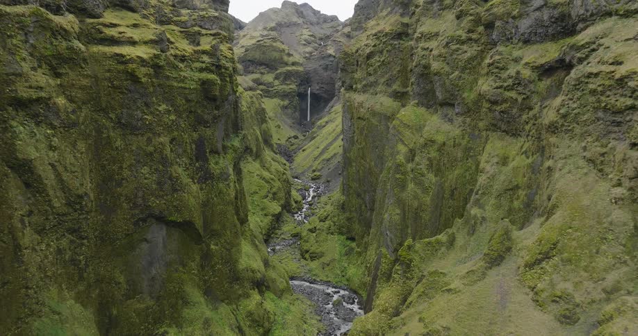An epic aerial view slowly flies through a lush green mossy canyon towards a beautiful waterfall in Iceland