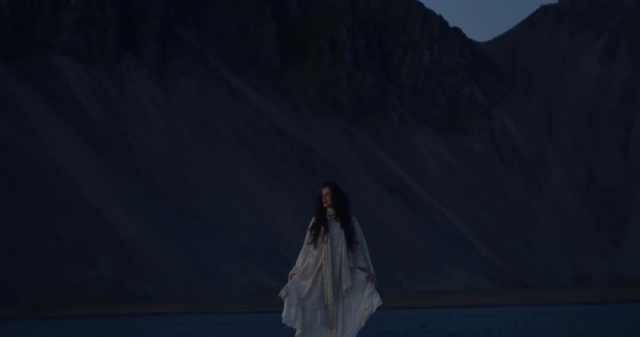 A mysterious woman in a flowing white vintage dress stands pensively in a dramatic, dark mountain landscape at twilight