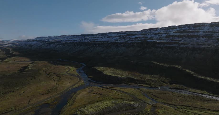 A stunning aerial view captures the vast, green valley and winding river below a majestic, snow-capped mesa under a blue sky