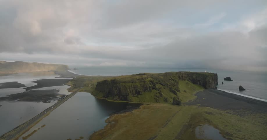 An epic aerial shot reveals the Dyrhólaey peninsula in Iceland, showcasing the lighthouse, green cliffs, black sand beaches, and surrounding ocean on a cloudy day