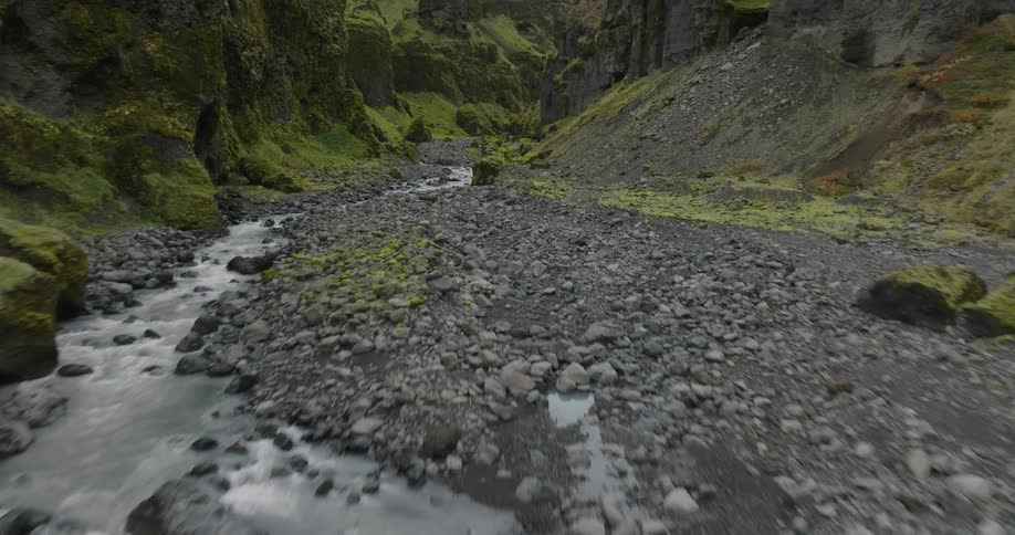 Low aerial view flying up a rocky river flowing through a lush green, moss-covered canyon in Iceland