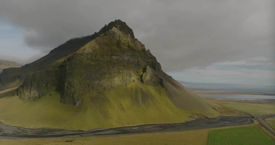 An epic aerial view slowly approaches a majestic, sunlit mountain peak towering over Icelandic farmlands and a glacial river