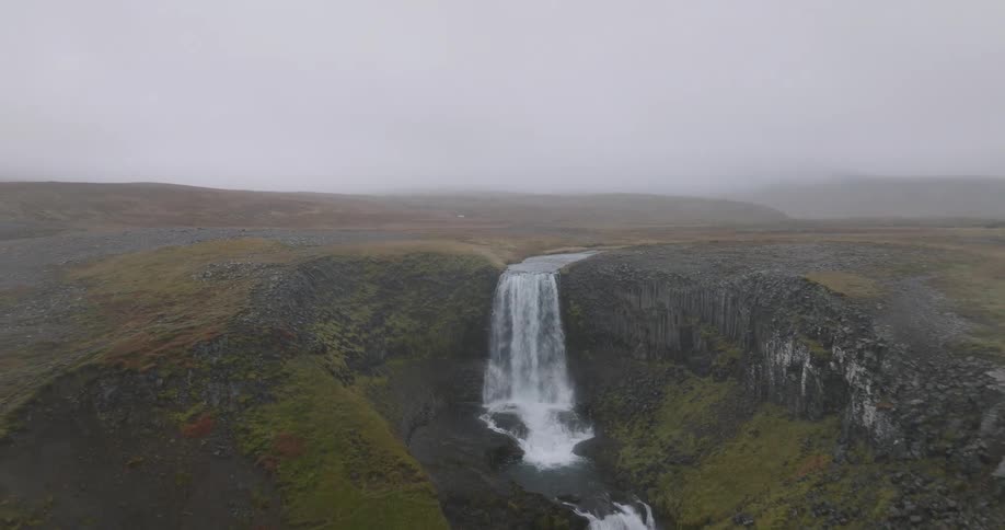Dramatic aerial footage pulls back from a powerful waterfall to reveal a vast, moody, and foggy Icelandic landscape with a winding river