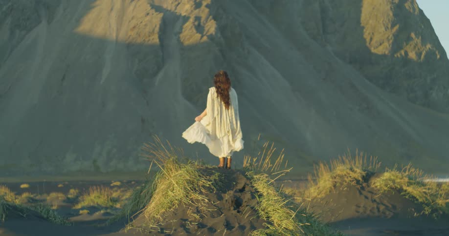 A woman in a flowing white dress stands on a black sand dune looking towards a majestic mountain at golden hour