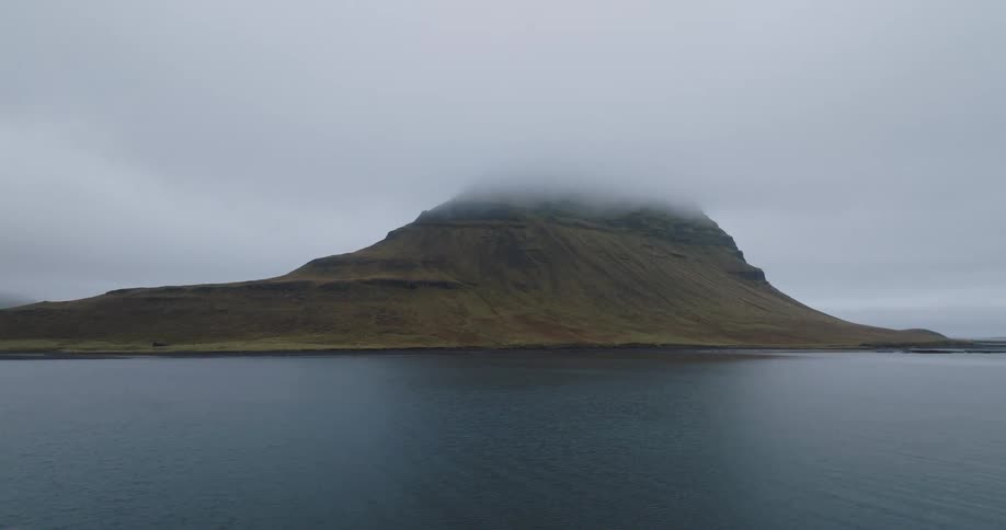 A wide, static shot of the iconic Kirkjufell mountain in Iceland, its peak shrouded in clouds, overlooking a calm body of water on an overcast day