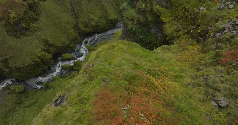 An aerial shot flies over a woman in a flowing dress sitting on a rocky cliff edge, revealing a majestic green canyon with a river running through it