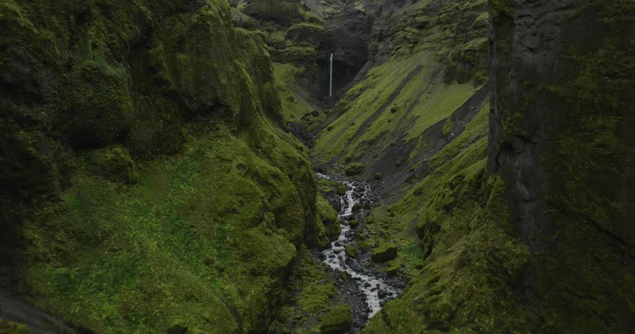 A slow aerial flight through a deep, moss-covered canyon with a river flowing towards a distant waterfall in Iceland