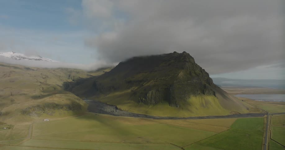 A majestic, moss-covered Icelandic mountain shrouded in clouds towers over a vast green valley as the camera slowly reveals the epic landscape