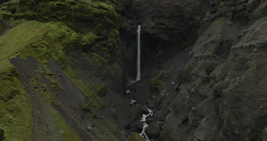A slow aerial approach through a dramatic moss-covered ravine towards a striking waterfall in the Icelandic highlands