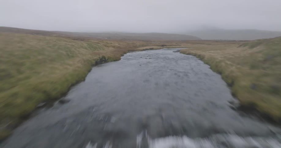 An aerial view soars up a fast-flowing river and over the edge of a stunning waterfall surrounded by basalt columns in a misty, rugged landscape