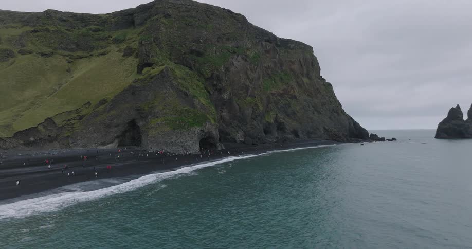 An aerial view pulls back from the famous Reynisfjara black sand beach in Iceland, revealing tourists exploring sea caves and the iconic sea stacks offshore