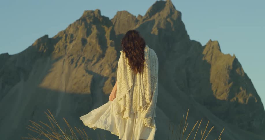 A woman in a flowing white dress and lace shawl stands before a majestic mountain at golden hour in slow motion