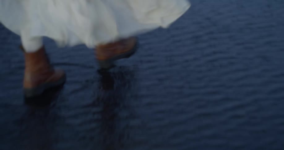 From behind, a woman in a flowing white dress and brown boots walks through shallow water on a black sand beach, with epic, dark mountains in the distance